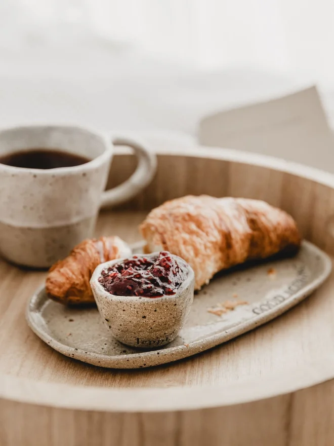 A Ceramic jam pot with a natural scalloped rim, filled with jam next to a croissant and mug of coffee