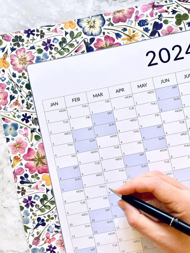 A Hand Holding a Pen Ready to Write on a Floral Yearly Wall Planner - Colourful Pressed Flowers and Leaves make up the Design