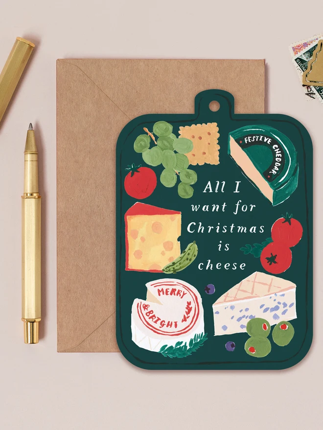 Dark green Cheeseboard Christmas card shaped like a board of cheeses, grapes and tomatoes, lettered 'All I want for Christmas is cheese', on a kraft envelope beside a gold pen