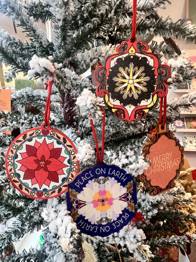 A set of four multi coloured wooden tree decorations are hanging by red ribbons on a fake white christmas tree with other decorations. 