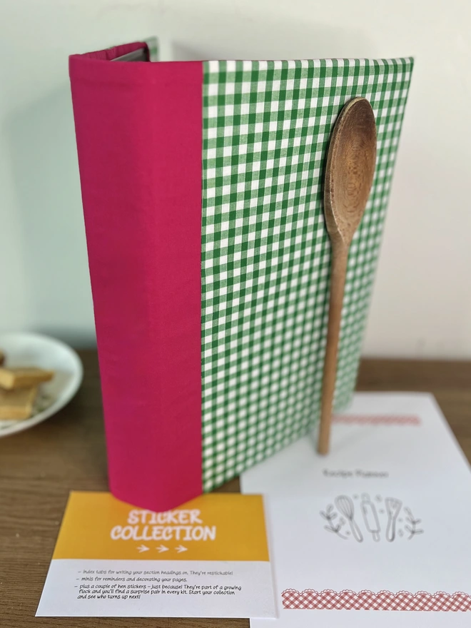 Green gingham binder with plain (non-embroidered) pink spine, standing upright on wooden table with recipe template pages & ‘Sticker Collection’ envelope beside it & a wooden spoon propped against the folder. Home-baked squares on plate in background.