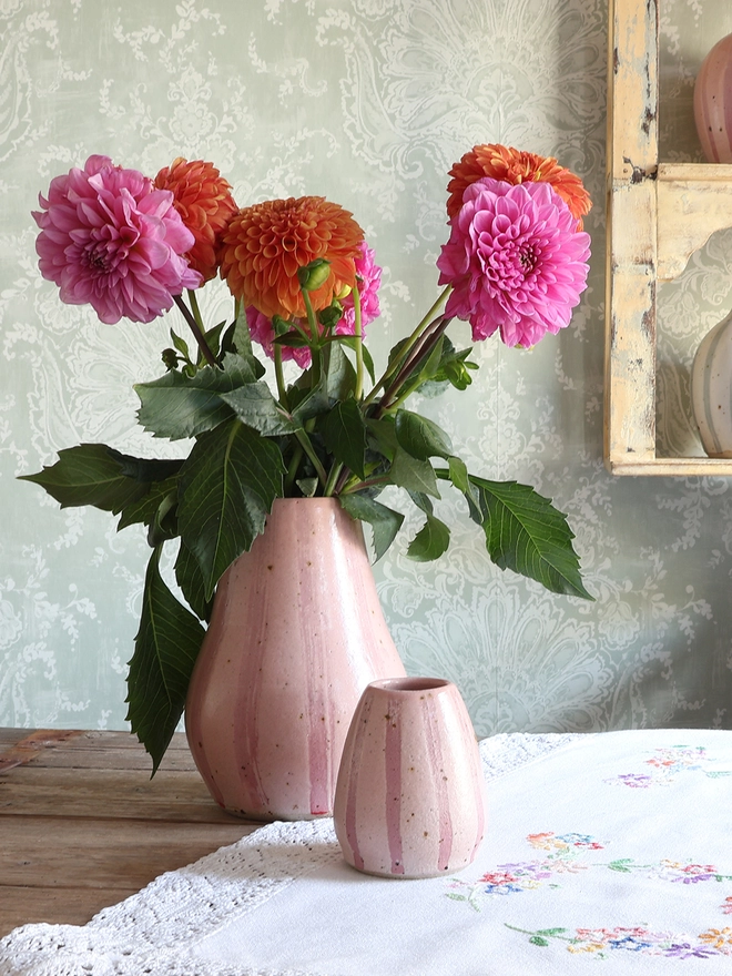 Handmade Ceramic Stoneware Large Vase in Pink Stripe filled with a bouquet of orange and pink flowers sat on a wooden surface covered in a vintage tablecloth.  A Small Handmade Ceramic Stoneware Vase in the same style sits in front of it. 