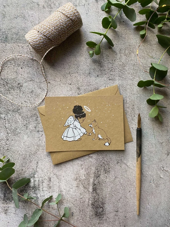 a Christmas postcard featuring a story book illustration of a sweet afro Caribbean angel girl and a fox cub printed on a Kraft postcard and laid on a Kraft envelope