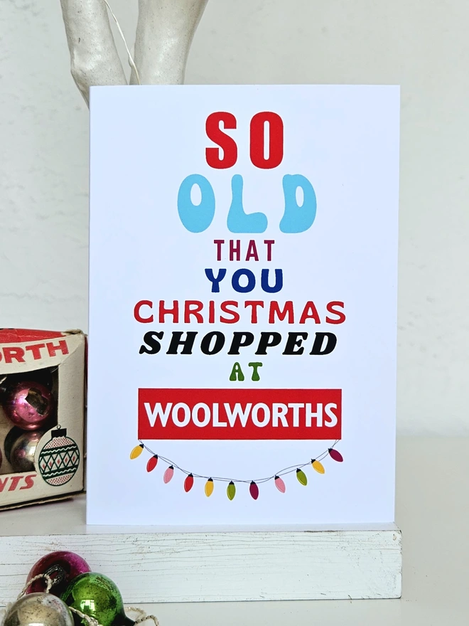 A photograph of the front of a humorous A6 greeting card illustrated using brightly coloured text and the retro shop logo. The text says so old you Christmas shopped at Woolworths with string lights