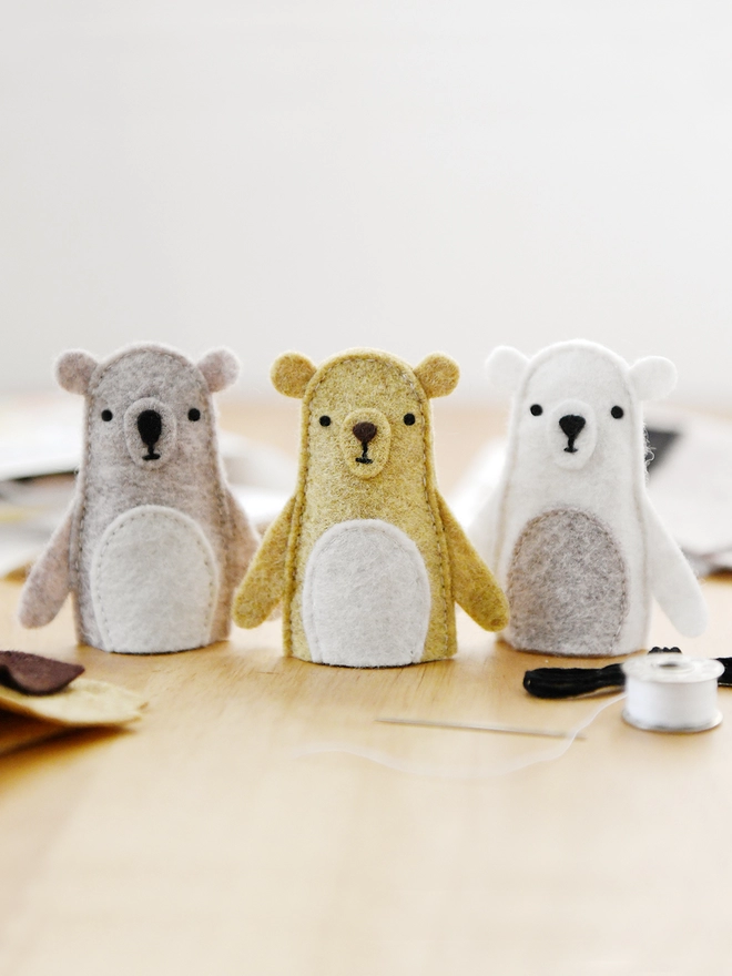 Three handmade felt bear finger puppets stand on a wooden desk with various craft kit materials beside them.
