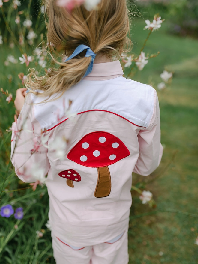 A girl stands in a garden wearing a pink shirt with a toadstool applique