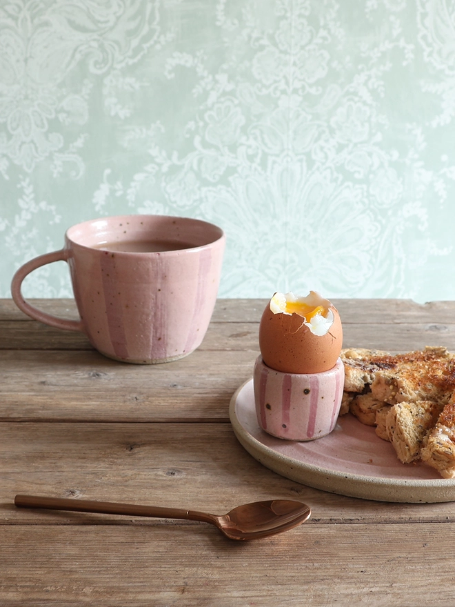 A pink-striped ceramic egg cup sits on a plate with buttered toast alongside a brass spoon and a matching mug on a wooden table.