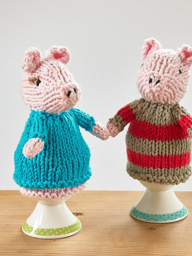 Egg Cosy Pigs Table Decoration (Set Of 2), a pair of colourful pig cosies sitting on a wooden surface. 