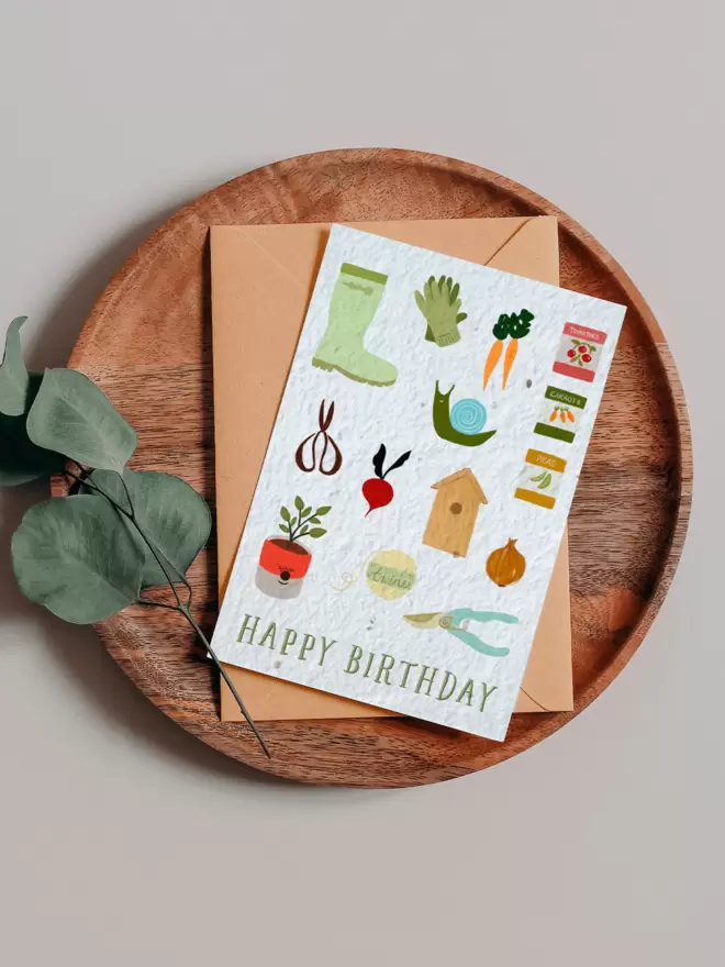 Happy Birthday Seeded Card with Gardening Illustrations including wellies, gloves, carrots, bird box, scissors, plant pot, onion, radish, seed packets, a snail and string on a wooden tray next to a Eucalyptus branch
