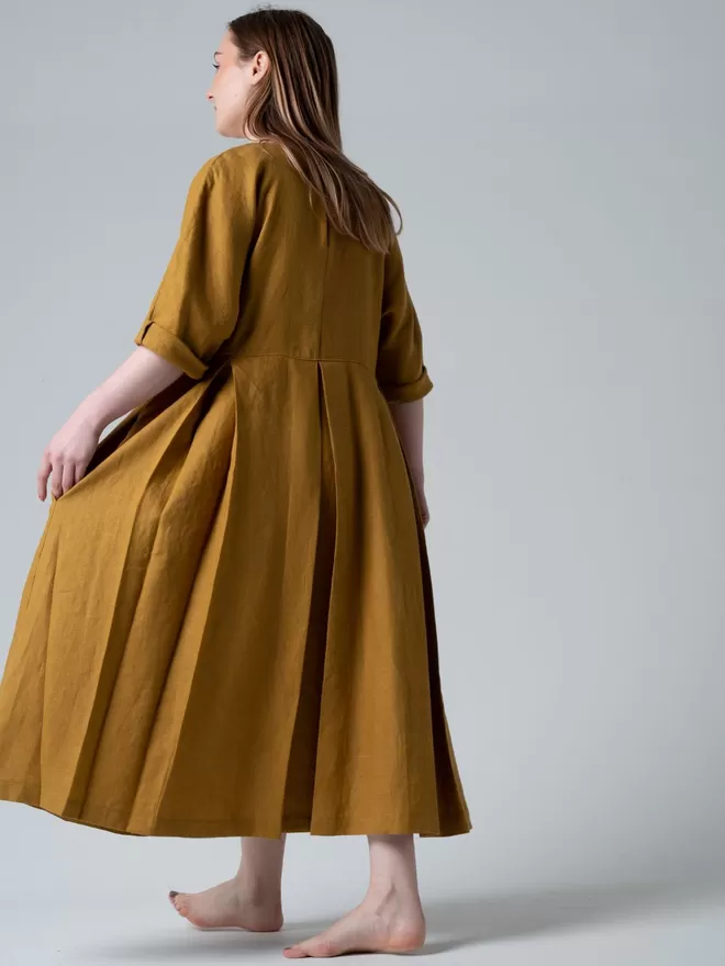 Midi length  luxury linen dress in golden colour. Deep pleated skirt with elbow length sleeves.  Studio back view.