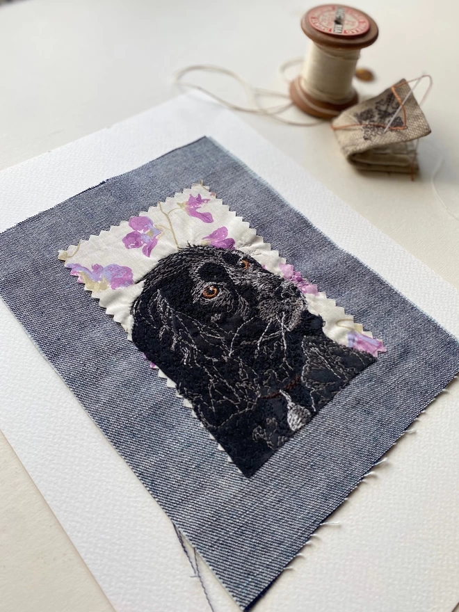embroidered pet portrait of a black dog, unframed, on a desk with a sewing tools