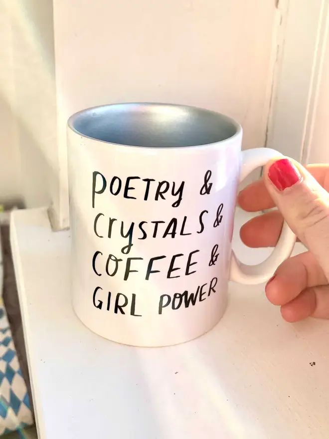 Holding mug which reads 'poetry & crystals & coffee & girl power' mug is silver inside