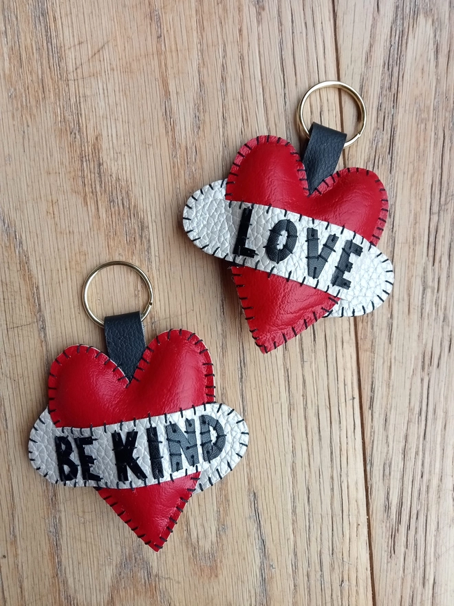 Two red leatherette tattoo style keyrings, with black lettering across white scrolls. One says LOVE, the other, BE KIND