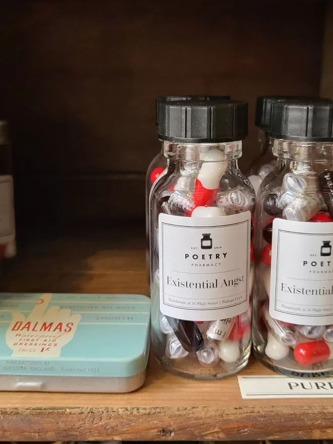 Two glass bottles of red, white and clear Existential Angst poetry pills on a wooden shelf, next to a vintage tin of sticking plasters