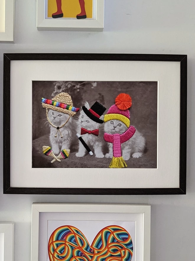 B&W print, 3 cats in embroidered sombrero, top hat and bobble hat, framed on wall
