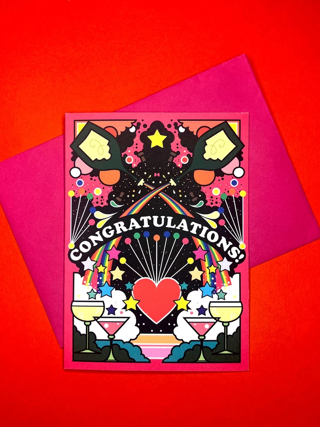 A vibrant pink greetings card with Congratulations written in white on a black background in an arch across the centre. The design is multi-coloured and features champagne flutes shooting from rainbows, hearts and stars. The card sits on a pink envelope on a red backdrop.