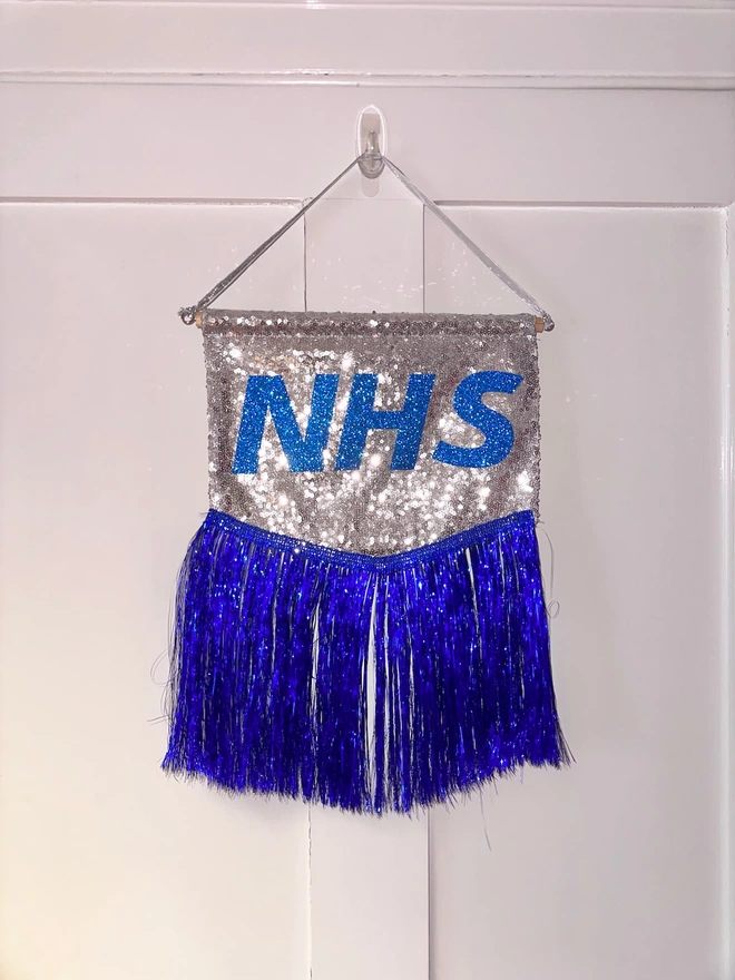 A tiny NHS banner hanging on a hook. The background is silver sequins and the NHS logo features in blue in the centre. It is trimmed with blue tinsel.