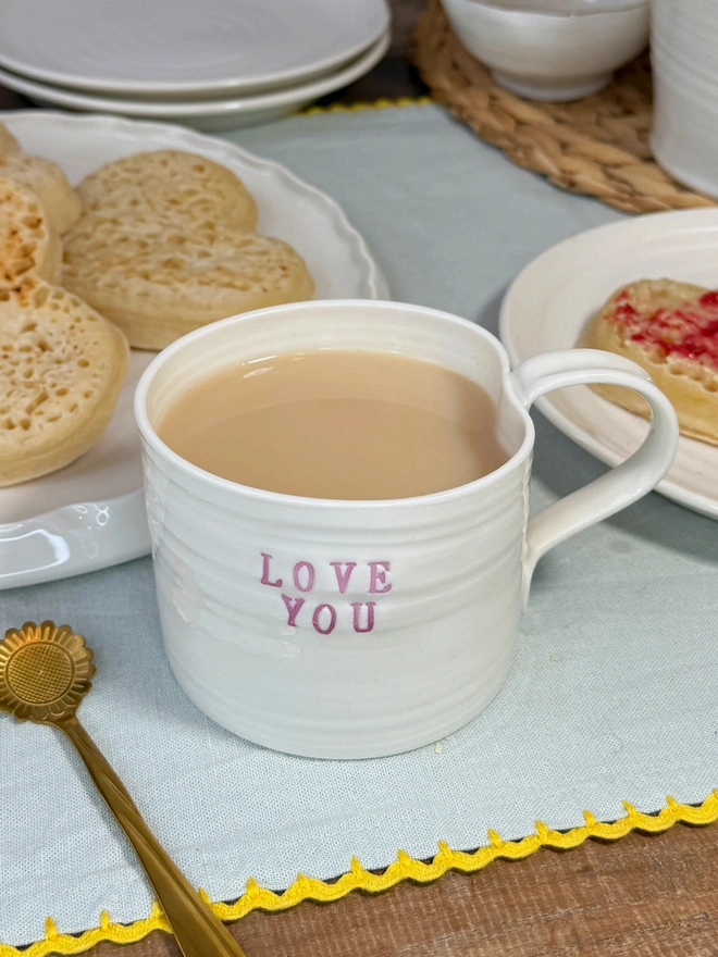hand thrown porcelain cream mug with pink lustre text on the side that says LOVE YOU. The mug is sat upon a sage table cloth amongst a tea time scene of heart shaped crumpets