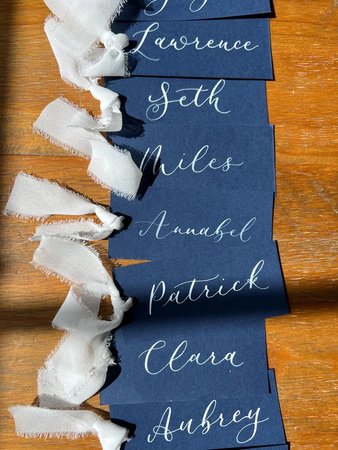 Personalised Calligraphy Wedding Navy Place Cards, navy place cards set together on a wooden table with white ribbons. 