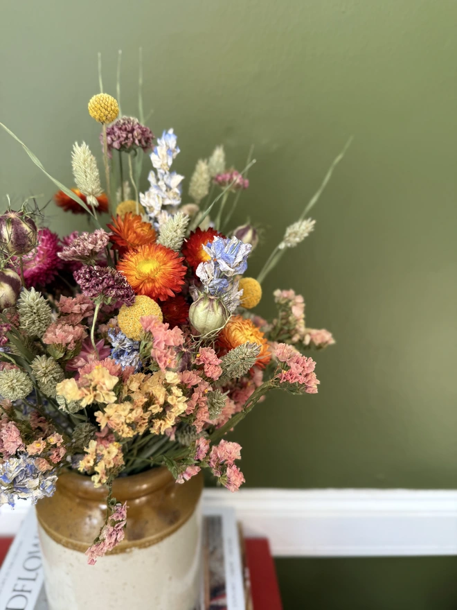 a dried flower bouquet of spring/summer colours in a victorian jam pot sitting on top of a stack of books and magazines against a green wall