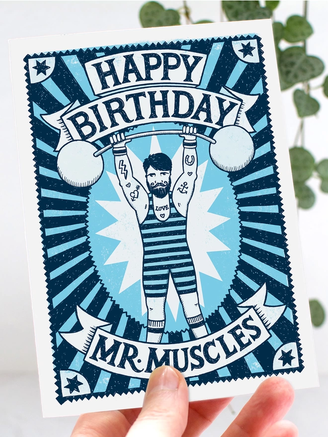 hand holding blue mr muscles muscle man birthday card with leaves in background