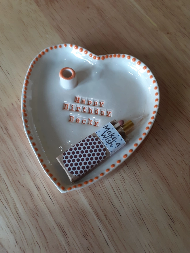 Handmade personalised ceramic heart dish with “Happy Birthday Becky” stamped into the clay, finished with an orange dotted rim and supplied with a gold birthday candle, two matches, and match striker sleeve.