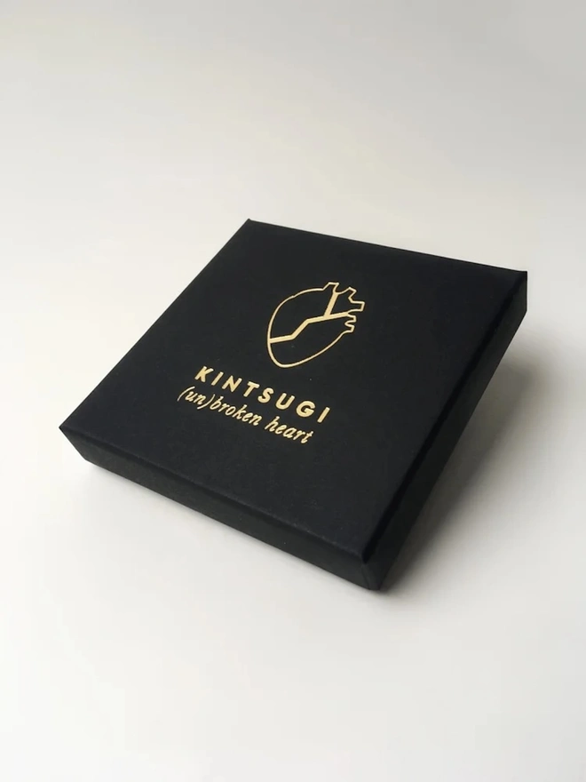 (Un)Broken Heart Pin, a black box with a card explaining the white and gold heart pin. 