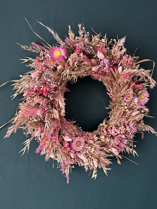 Pink Dried Floral Wreath. a circular wreath made of pink dried flowers hung up on a forest green wall.