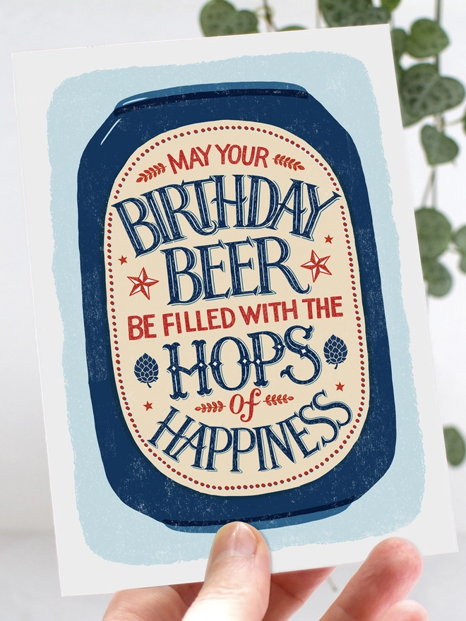 Illustrated beer can with hand lettered beer hops quote on a blue background held in a hand with leaves