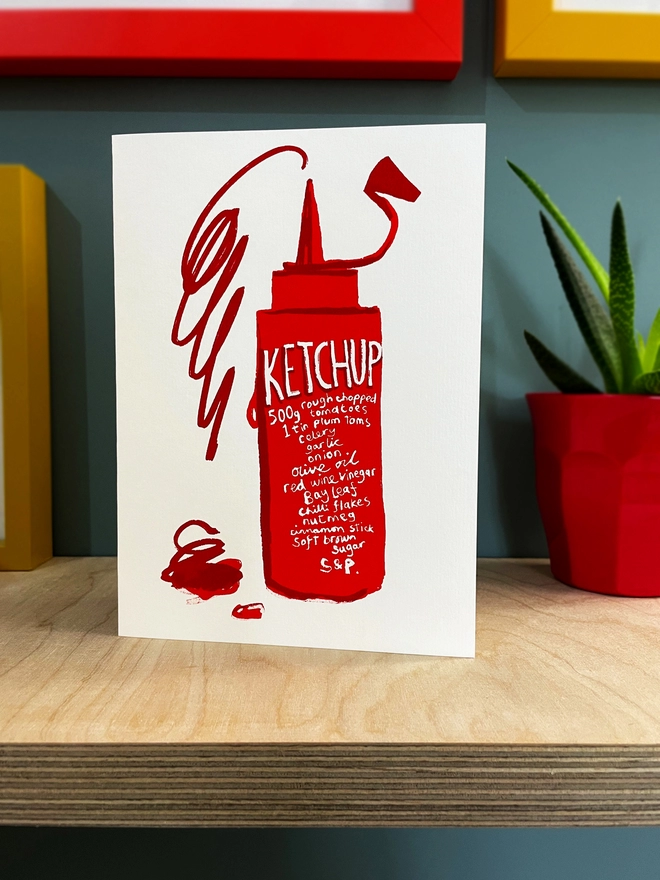 Ketchup recipe is written on a squeezy red bottle, with a scribbled squirt of ketchup jumping out the top - all on a white card stood on a plywood shelf. Bits of frames are shown around, and a plant to one side.