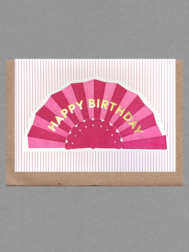 White card with pink fan with gold text reading 'Happy Birthday' on pink striped background with brown envelope behind