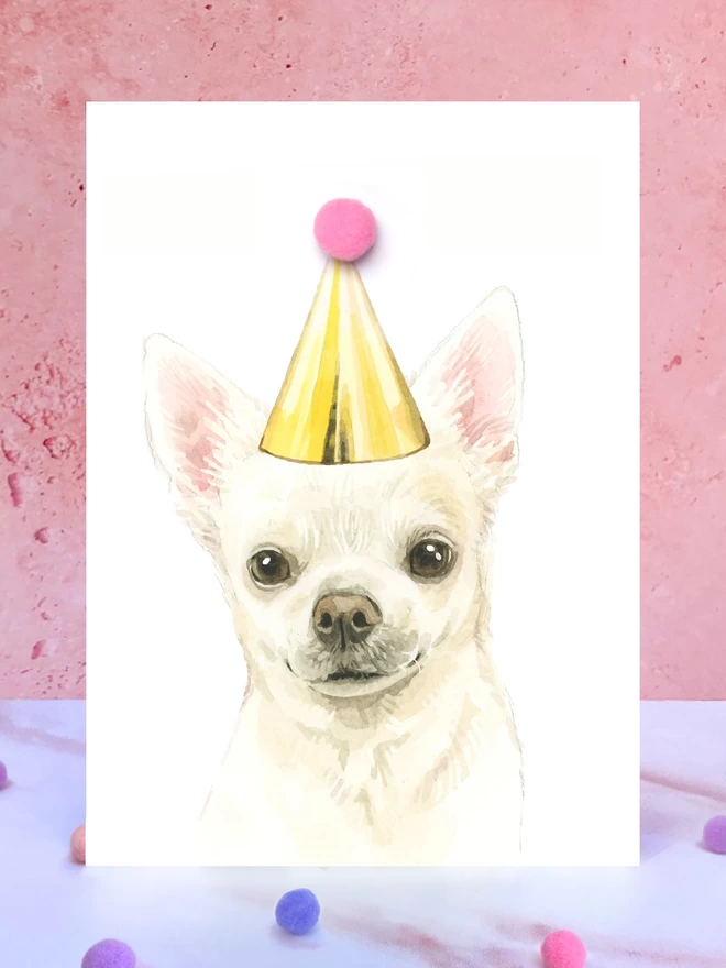 A greeting card featuring a hand painted design of a chihuahua, stood upright on a marble surface surrounded by pompoms. 