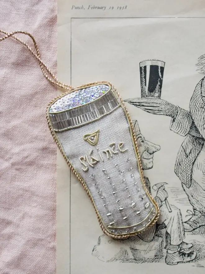 embroidered linen irish stout decoration. pale beige irish stout pint hanging ornament photographed on pink fabric background.