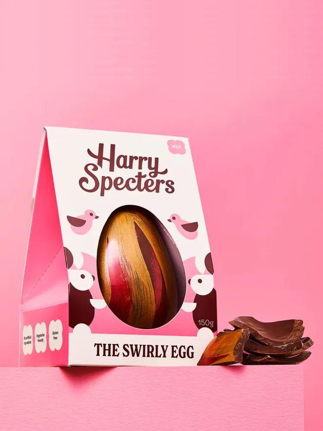 A milk chocolate Easter egg decorated with red and gold cocoa butter, next to a stack of milk chocolate Easter egg pieces