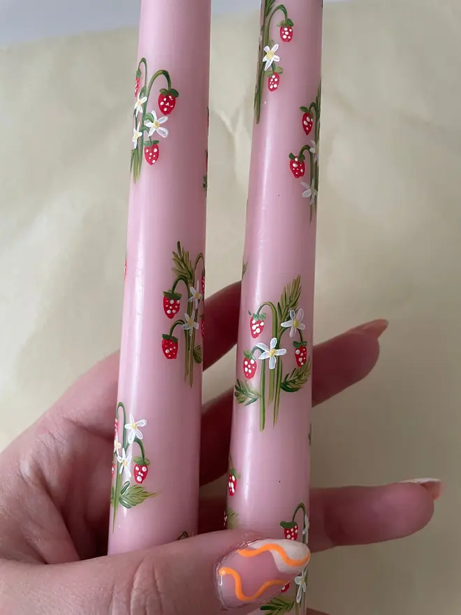 Wild Strawberry Pink Hand Painted Candle, a woman holding a pair of pink candles against a white backdrop. Their floral design wraps around them. 