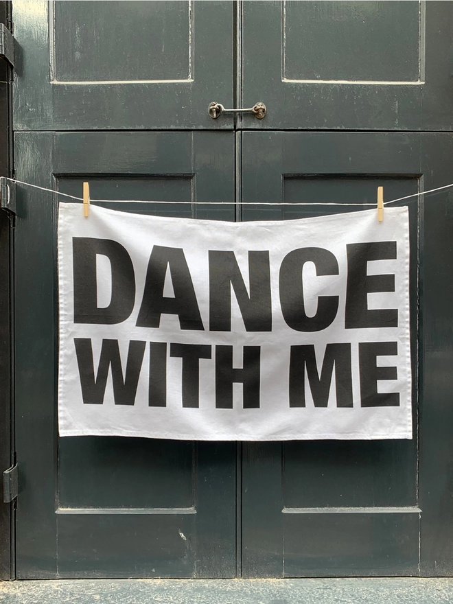 London Drying Dance With Me black text on white tea towel hanging washing line style in front of grey shutters