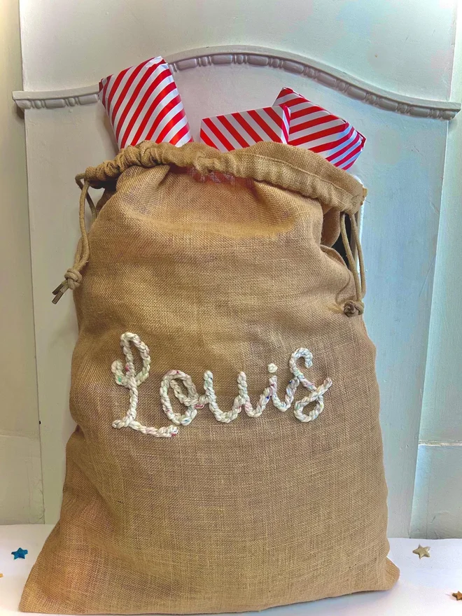 Personalised Christmas Sacks Louis, a Christmas sack sitting in front of a fireplace. It has its own personalisation and is filled with presents. 