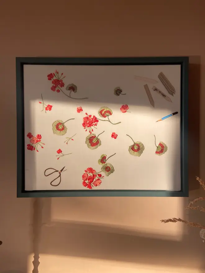 Framed embroidered geranium artwork with scattered red flowers, green leaves, scissors, pencil and fabric swatches on natural linen.