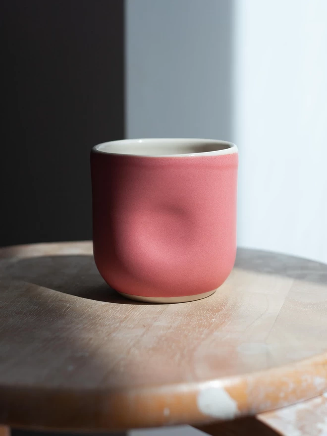 Close up of a pink ceramic cup with fingerprint dimples on either side, with a matte glaze, white wall in background