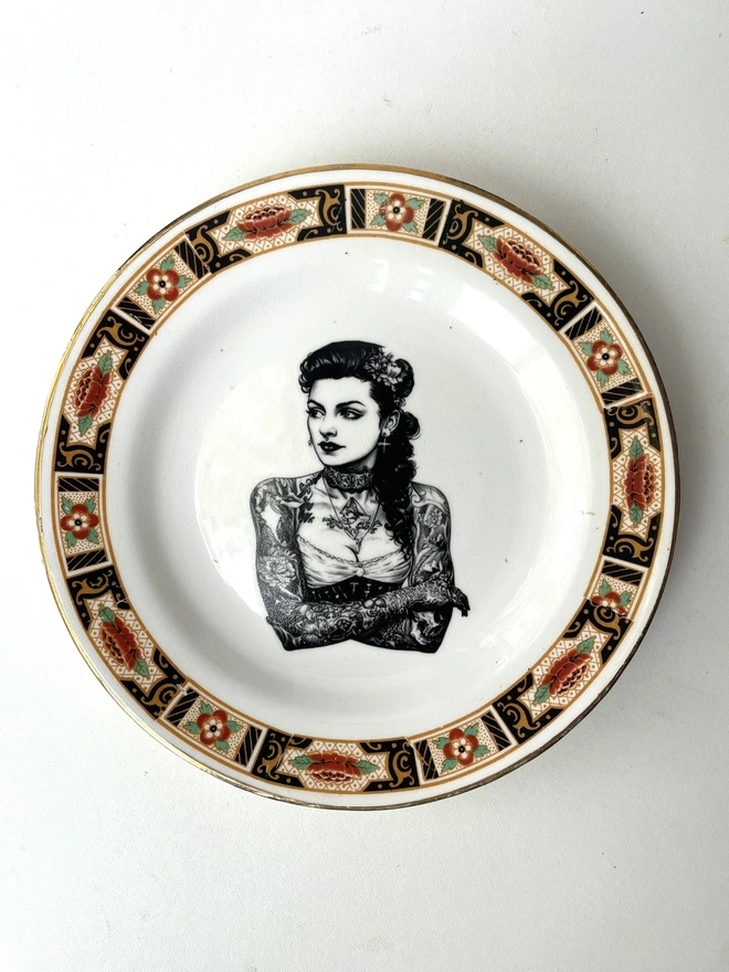 a beautifully ornate plate and in the centre is a vintage image of a beautiful tattooed london lady, she has ringlets and a choker and a tattooed ship on her chest