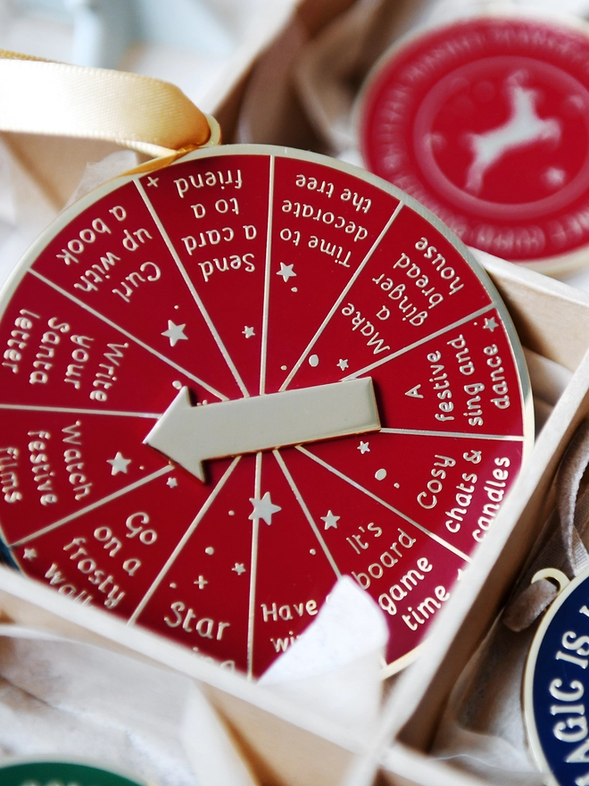 A deep red and gold Christmas decoration is tucked into a decorations box. It has 12 segments, each one with a different Christmas activity idea, and a golden arrow in the centre.