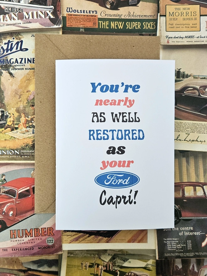 A birthday card that says you're nearly as well restored as your ford capri written in vintage style font on a white card. The colours of the words are blue, red and black.