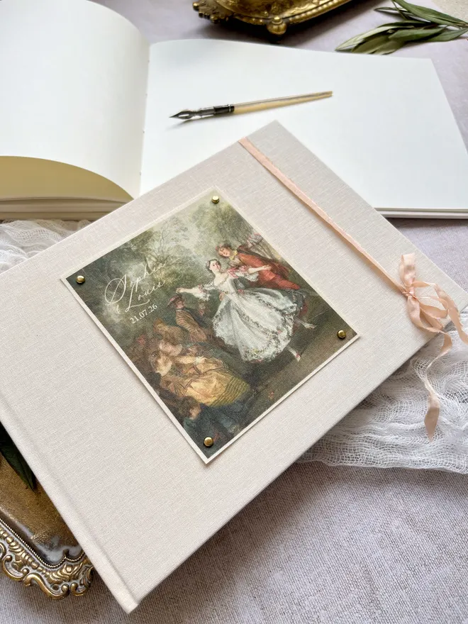 Luxury ivory linen guest book with a fabric printed image of dancers taken from an 18th century rococo work of art. A bow is added for a beautifully romantic finishing touch.