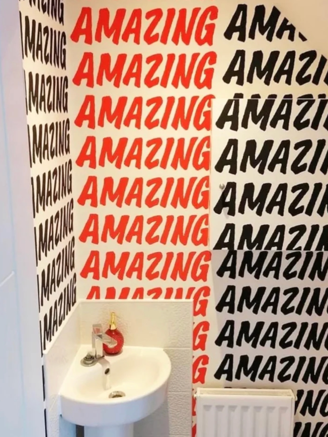 Amazing Wallpaper. A bathroom with a mixture of red and black strips of wallpaper which reads AMAZING, repeated in an all caps font.