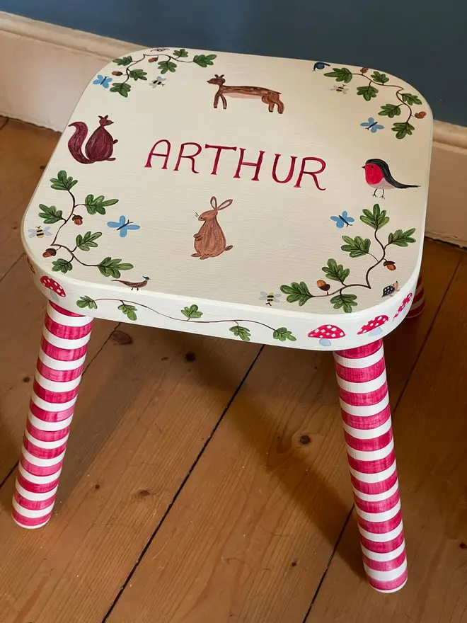 personalised hand-painted nursery stool. a small white stool with red and white striped legs, painted with a woodland scene and name