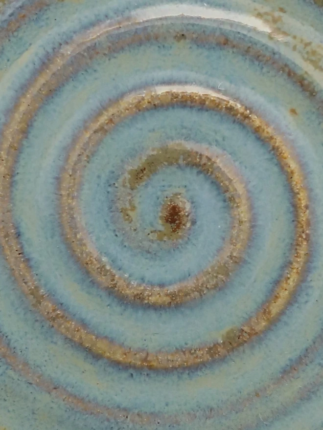 A close-up of a handmade ceramic glaze swatch in seafoam blue, showing a concentric swirl texture with warm sandy tones where the clay shows through