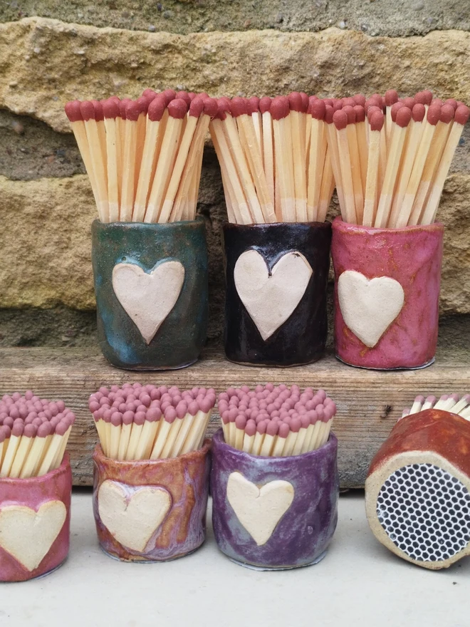 Heart Design Handmade Ceramic Match Holder & Strikers, a group of colourful match holders formed by two rows against a brick wall on a white surface. 
