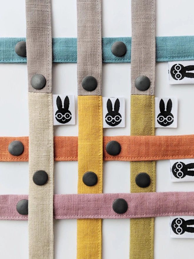 Handmade toy straps in soft neutral and pastel tones, featuring sturdy snap fastenings and small fabric tags with a bunny logo, neatly arranged in a row.