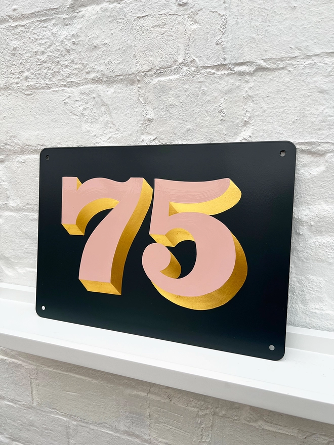 Hand painted house number against a white brick wall, number 75 in serif style in dusky pink and directionally burnished 23 carat gold leaf, on anthracite grey background. 