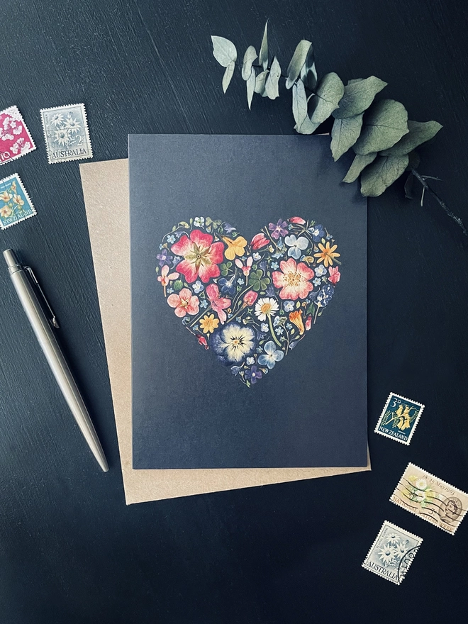 Inky Coloured Greetings Card with Digitally Printed Pressed Flower Heart Design - Brown Kraft Envelope - Dark Charcoal Coloured Desk - Colourful Flower Stamps, Silver Parker Pen, Dried Eucalyptus